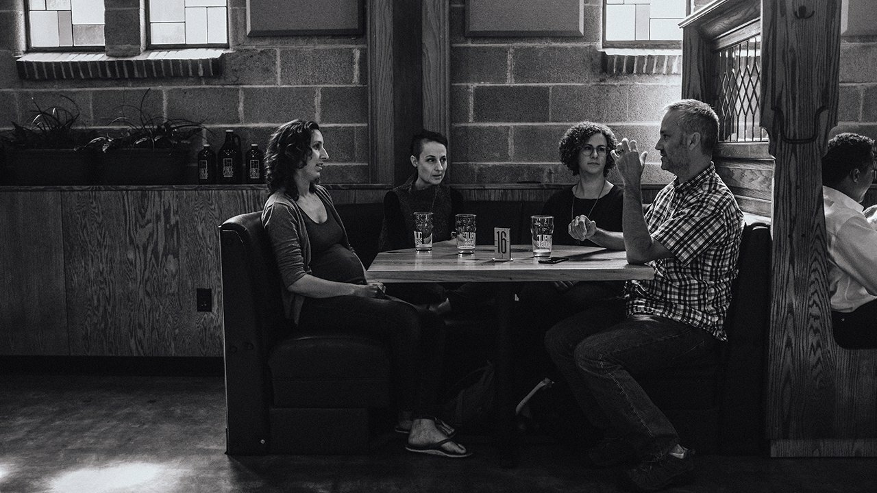 Two couples talking over pints in a dim brick-walled pub booth, black-and-white