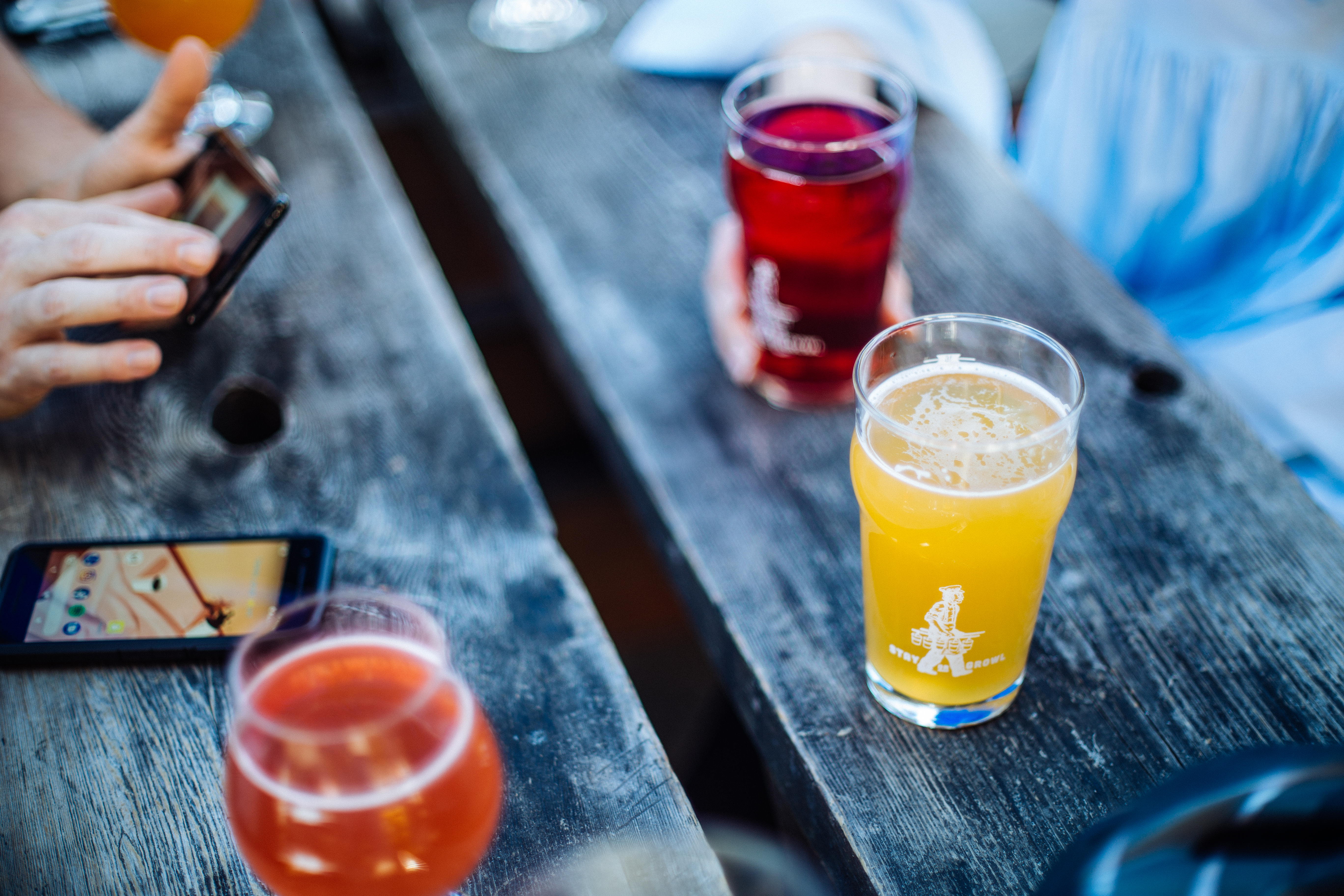 Hands holding colorful beers and phones on a weathered wood pub table