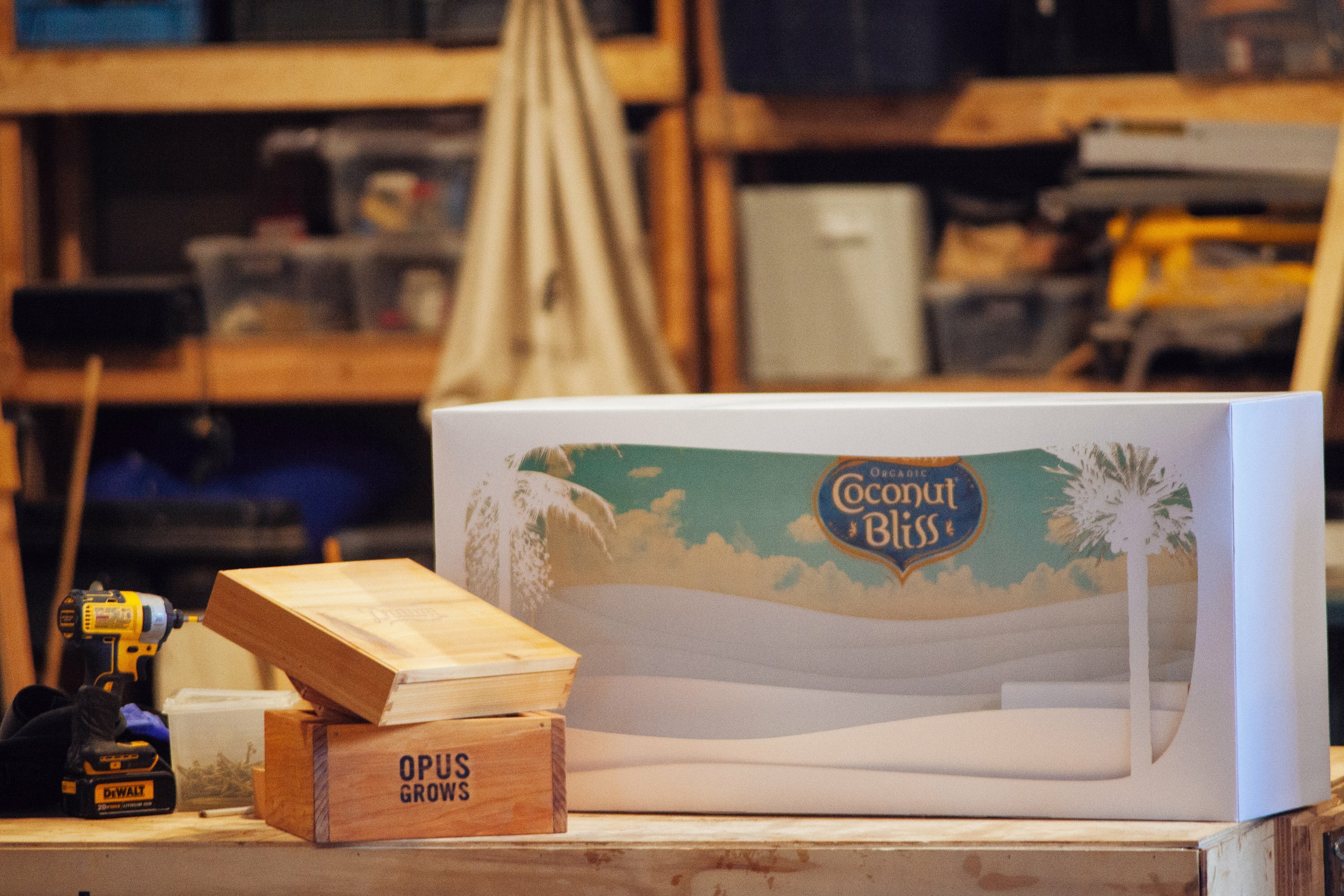 Coconut Bliss branded display box on a workbench beside an Opus Grows crate and drill in the workshop