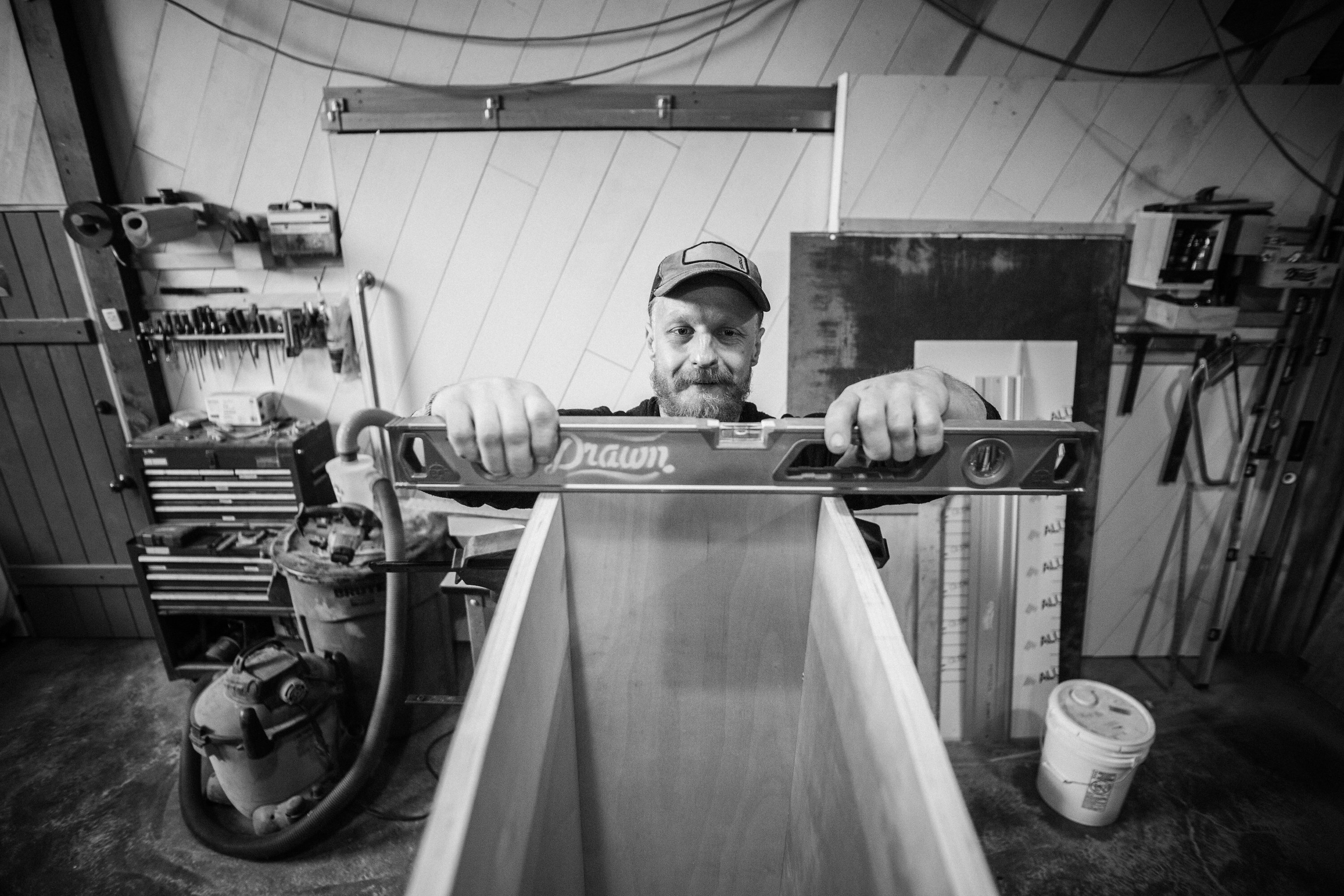 Maker in cap holding a Drawn level atop a wood form in the fabrication workshop, black and white