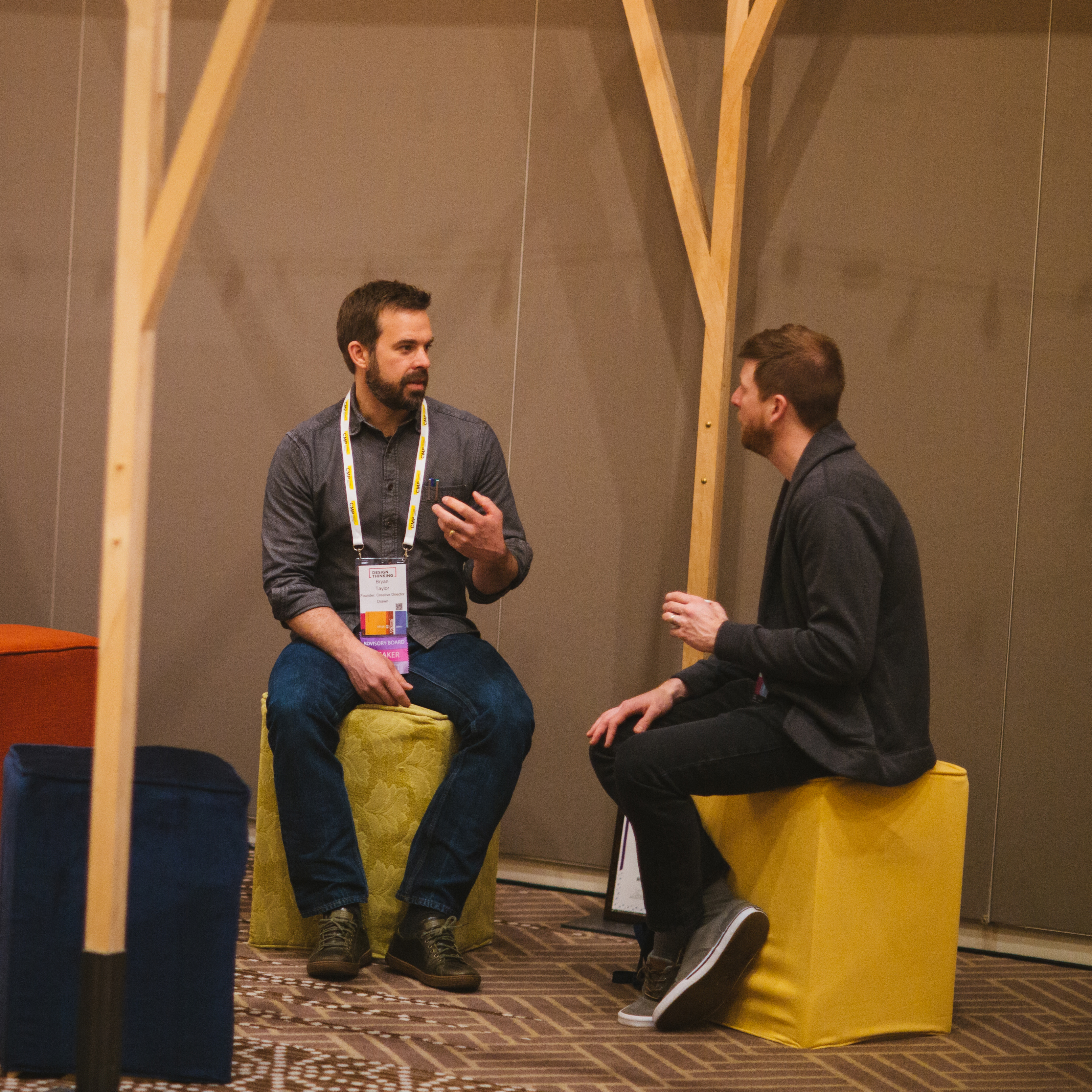 Two bearded men in conversation on yellow cube seats by a wooden frame at a design conference