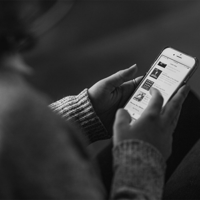 Person in a knit sweater browsing a podcast app on a phone, shown in black and white