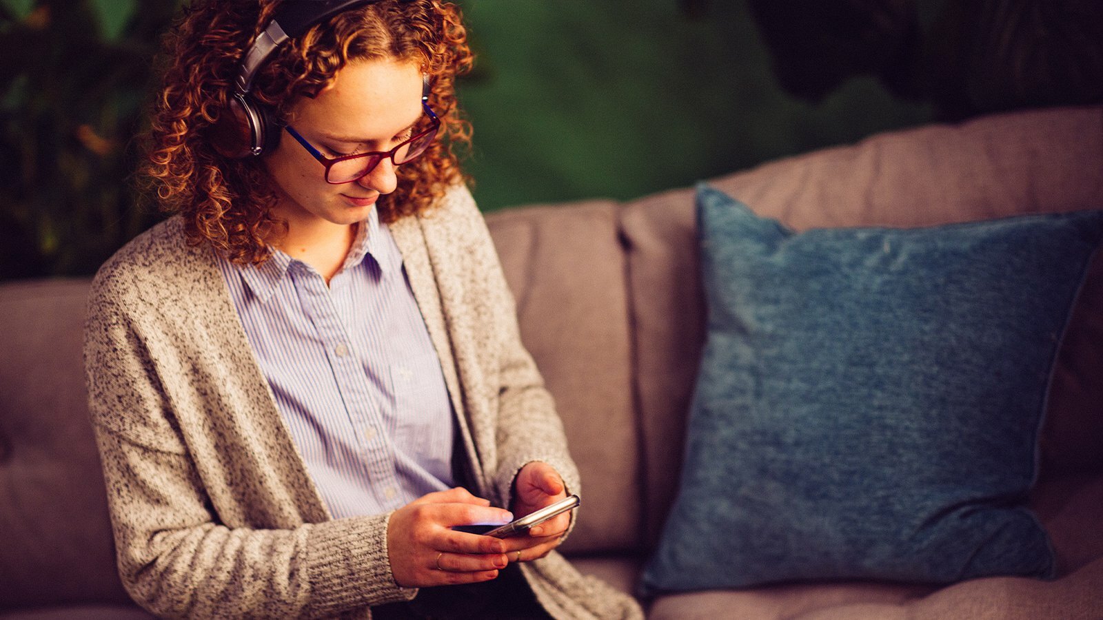 Woman in headphones and glasses using her phone while seated on a couch, listening to a podcast