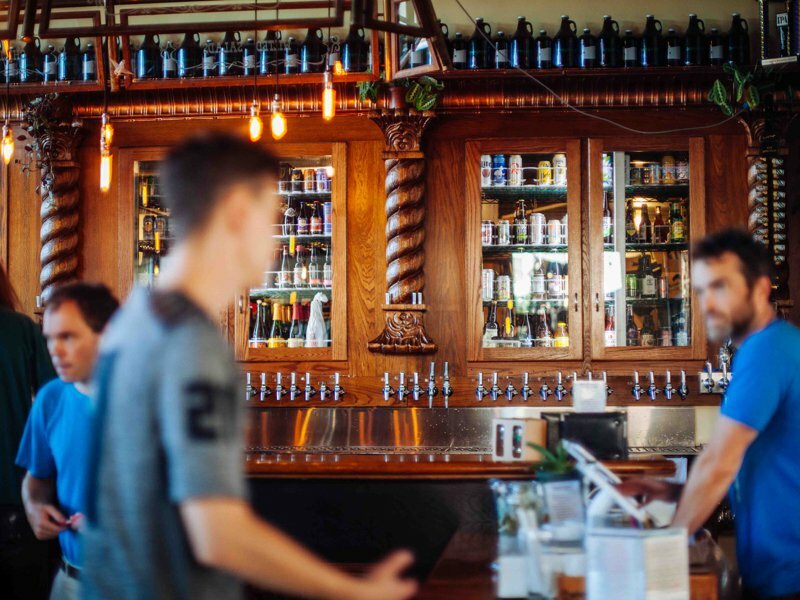 Carved wood back bar lined with growlers and bottles as staff and patrons move past taps