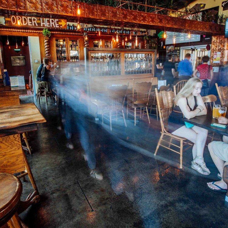 Patrons in a pub with pressed-tin ceiling and Order Here sign, figures blurred in motion