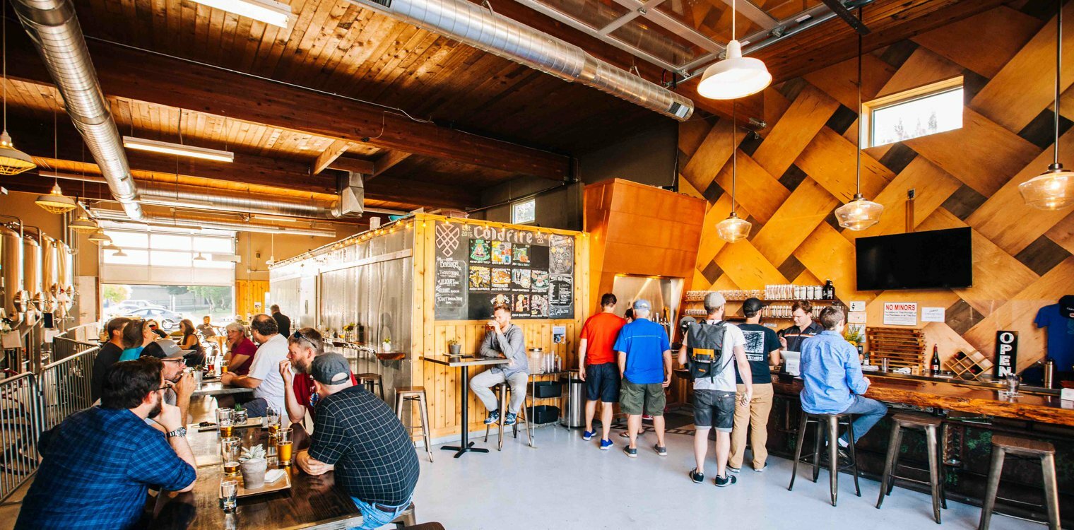 Sunlit brewery taproom with diamond-pattern wood wall, patrons ordering at the counter and seated at tables