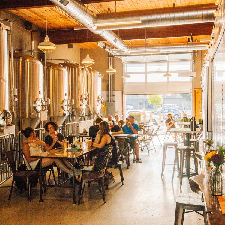 Brewery taproom with stainless fermentation tanks, exposed ducts and patrons drinking beer at communal tables