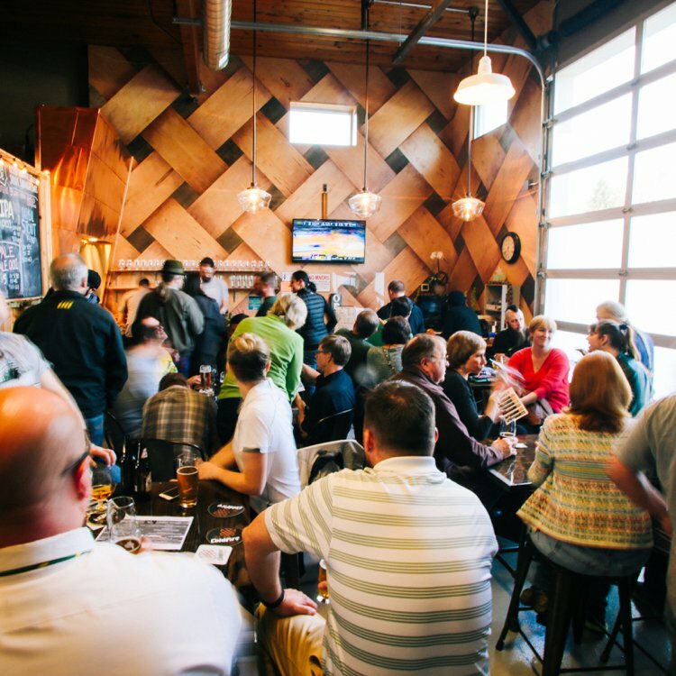 Crowded brewery taproom under a diamond wood wall and garage door, patrons drinking and watching a wall-mounted TV