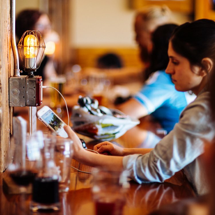 Patron charging a phone at a wood bar lit by a caged Edison bulb, other guests blurred behind
