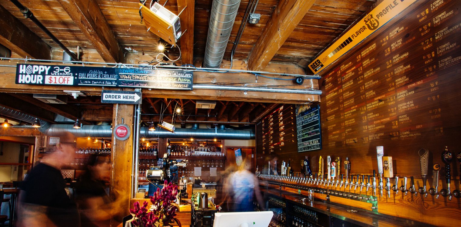 Brewpub bar with 'Hoppy Hour' and 'Order Here' signs, long row of beer taps and a wood tap list wall