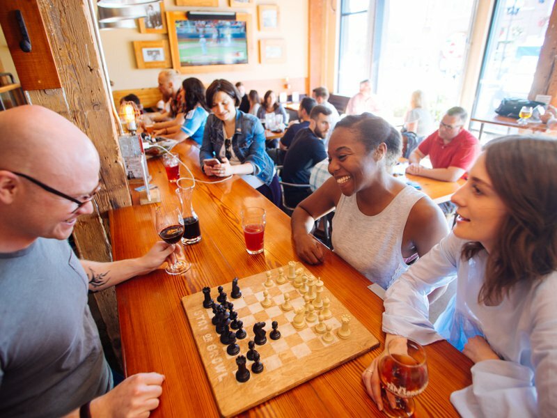 Friends laughing over a chess board and drinks at a communal wood bar in a busy brewpub