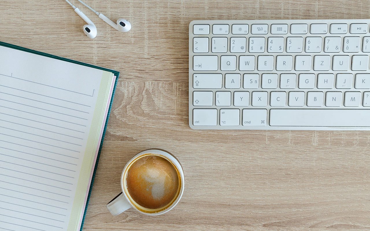 Flat-lay of an open lined notebook, white keyboard, earbuds and a cup of coffee on a wood desk