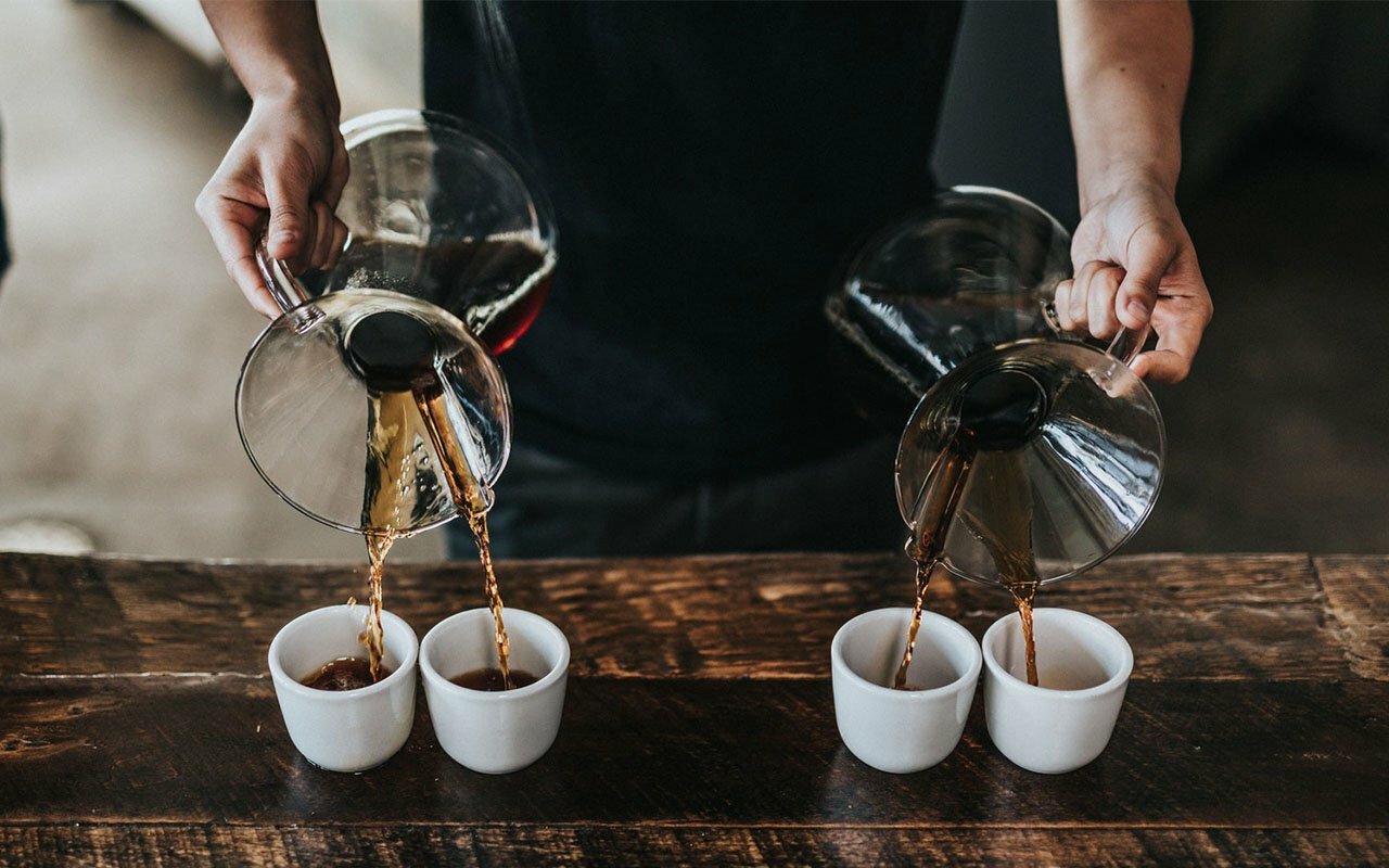 Barista pouring coffee from two Chemex carafes into four white cups on a wood table