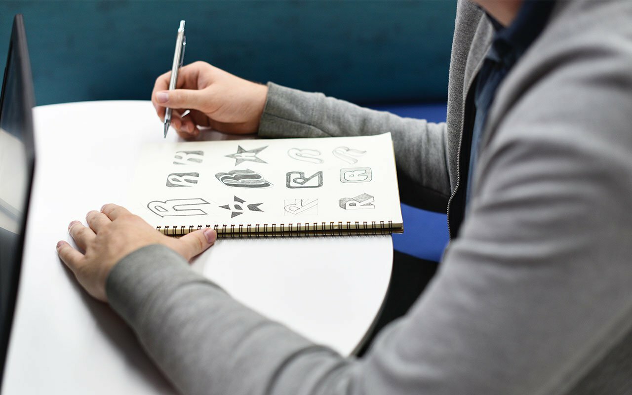 Person sketching rough logo concepts in a spiral notebook beside a laptop