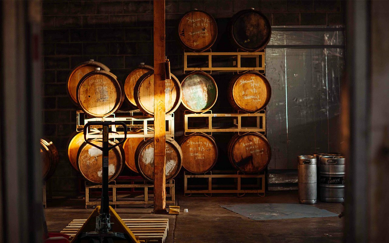 Oak barrels stacked on racks with metal kegs in a dim brewery or distillery cellar