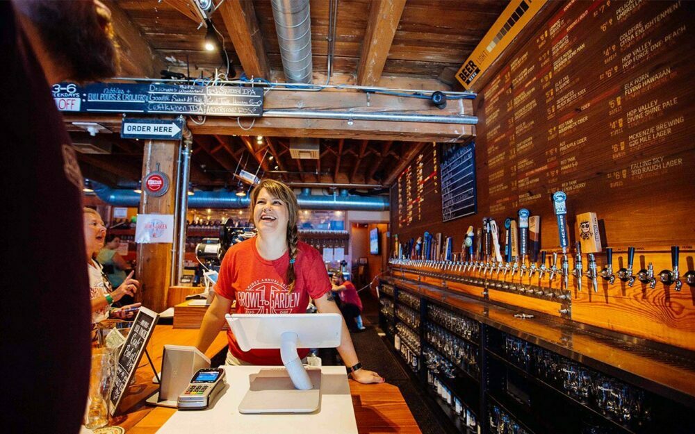 Smiling server at a brewery taproom counter lined with beer taps and chalkboard menus