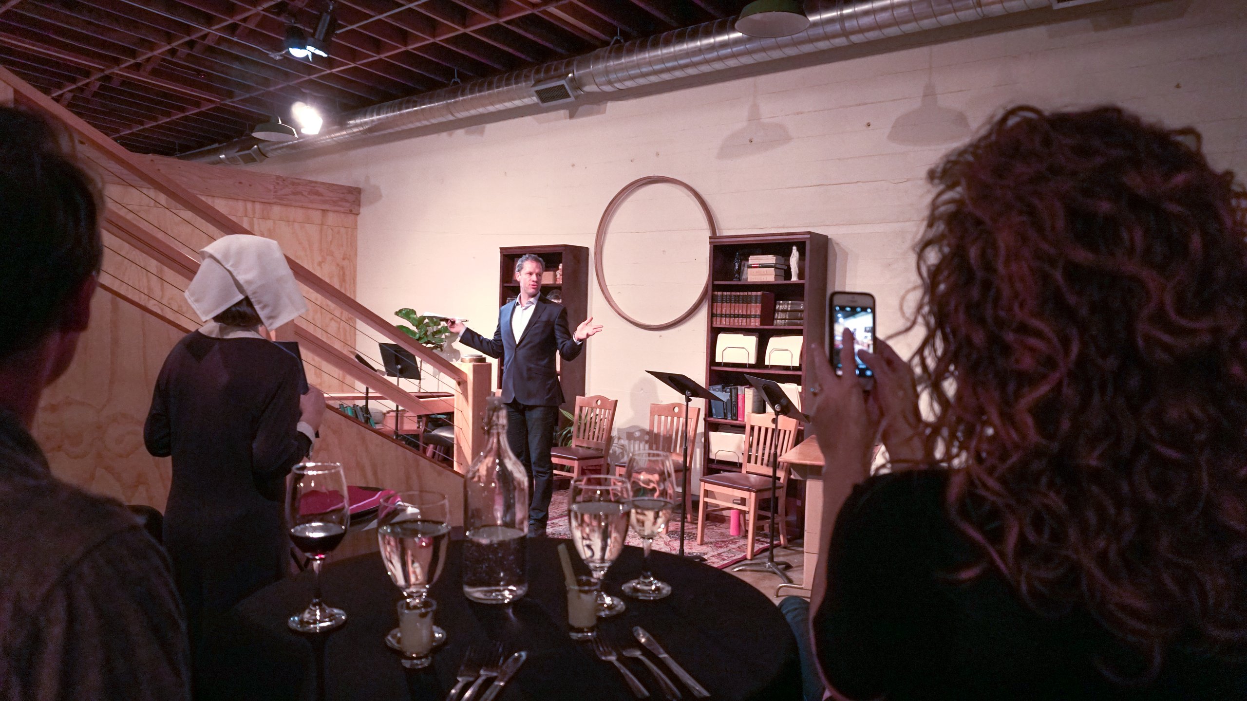 Man in a suit performing on a bookshelf-lined set while seated diners watch and film