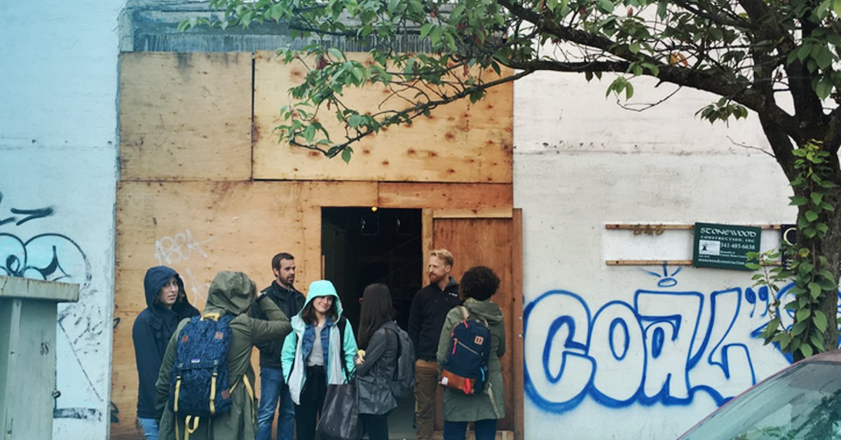 Group gathered at a plywood-fronted building entrance beside blue "COAL" graffiti and a tree