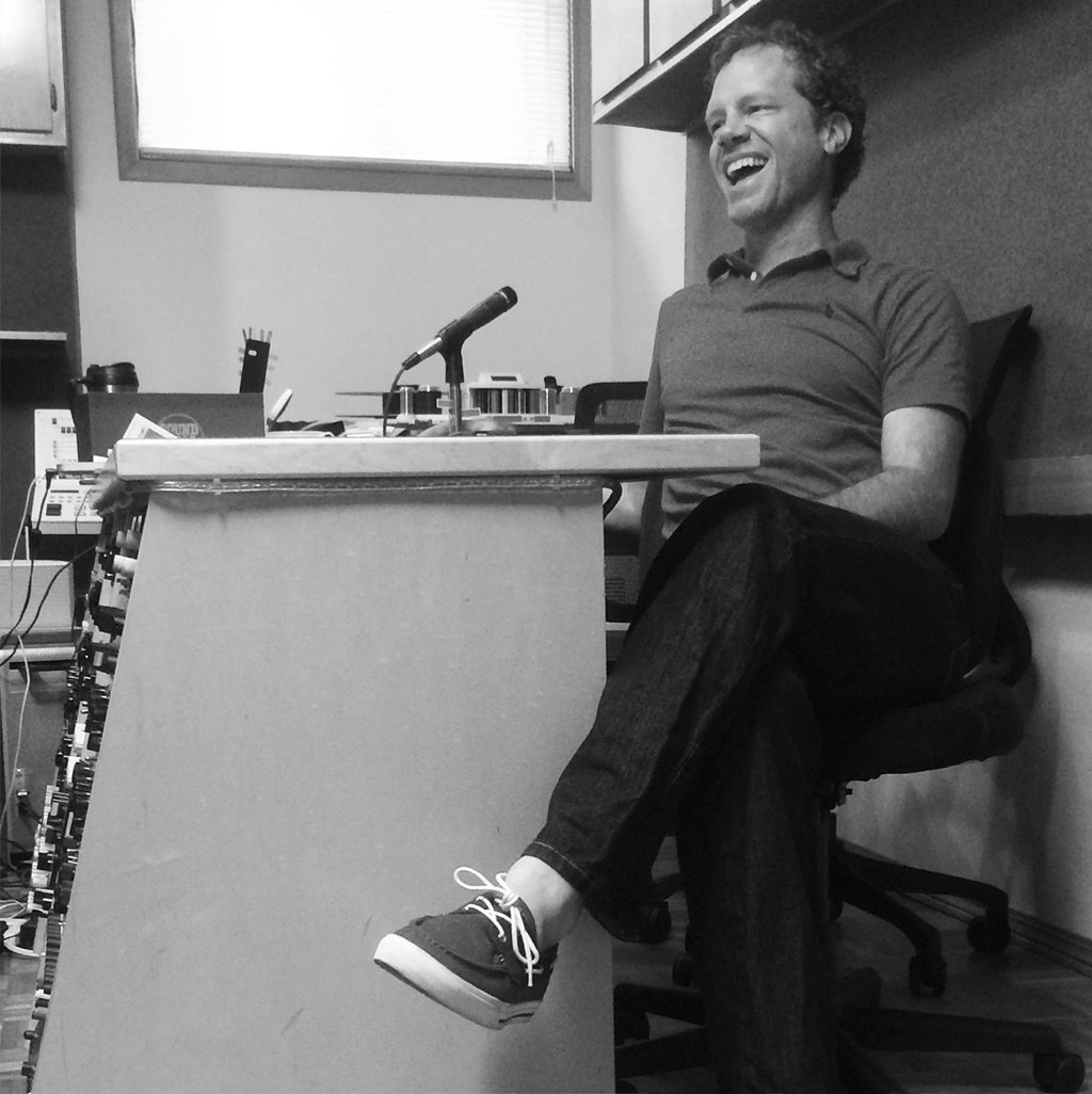 Black-and-white photo of a laughing man leaning back in an office chair, leg crossed