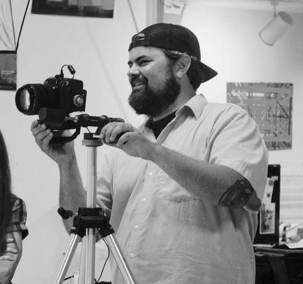 Black-and-white photo of a bearded man in a backwards cap filming with a tripod camera