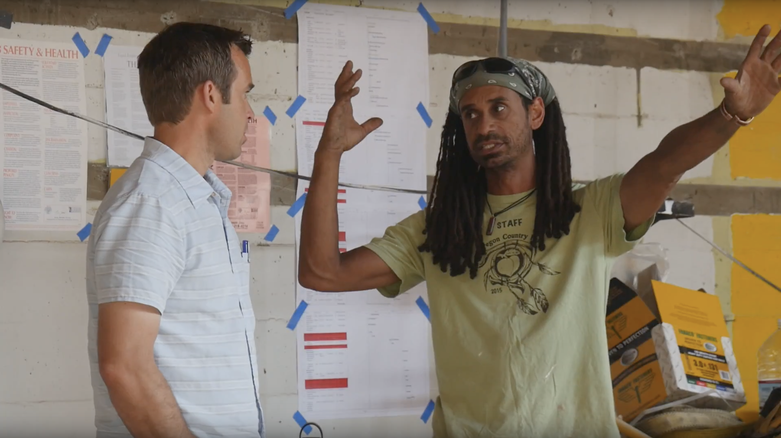 Two men talking in a concrete workshop, one in a bandana and STAFF tee gesturing