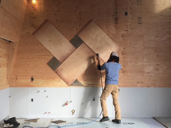 Person sanding angled plywood panels mounted on a plywood wall under construction