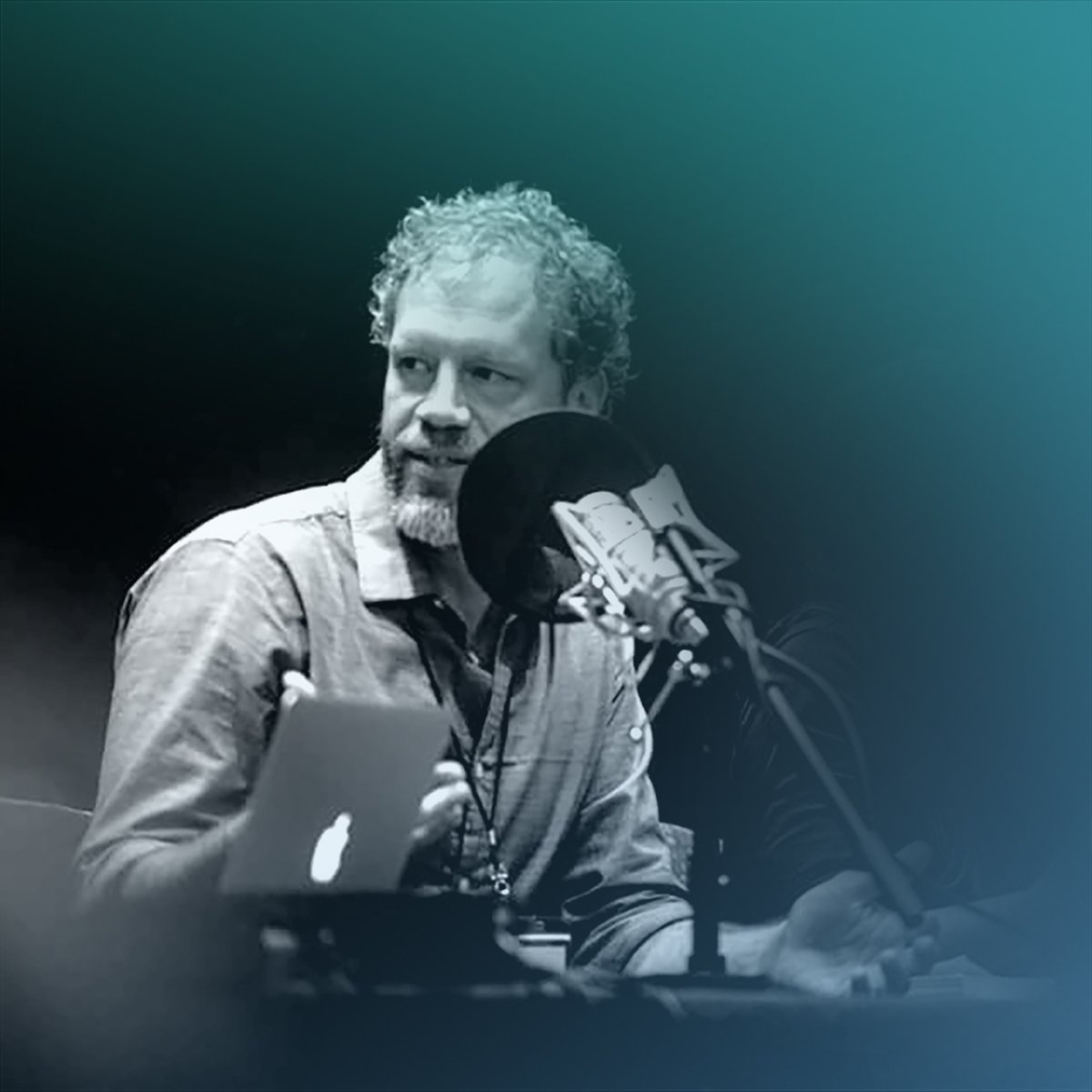 Teal-toned photo of a bearded man speaking into a microphone beside a laptop