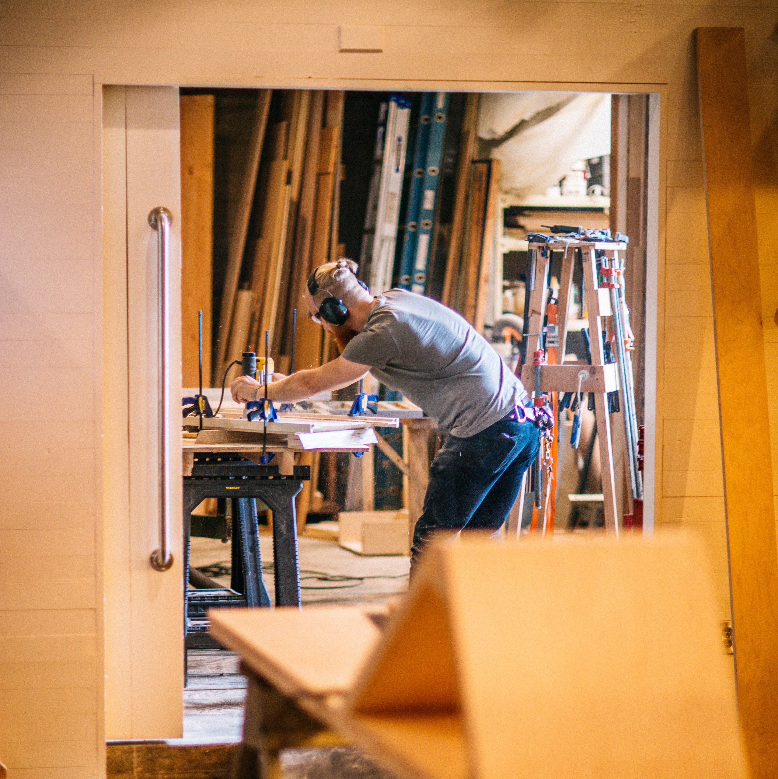 Man in ear protection working at a woodshop bench seen through a doorway