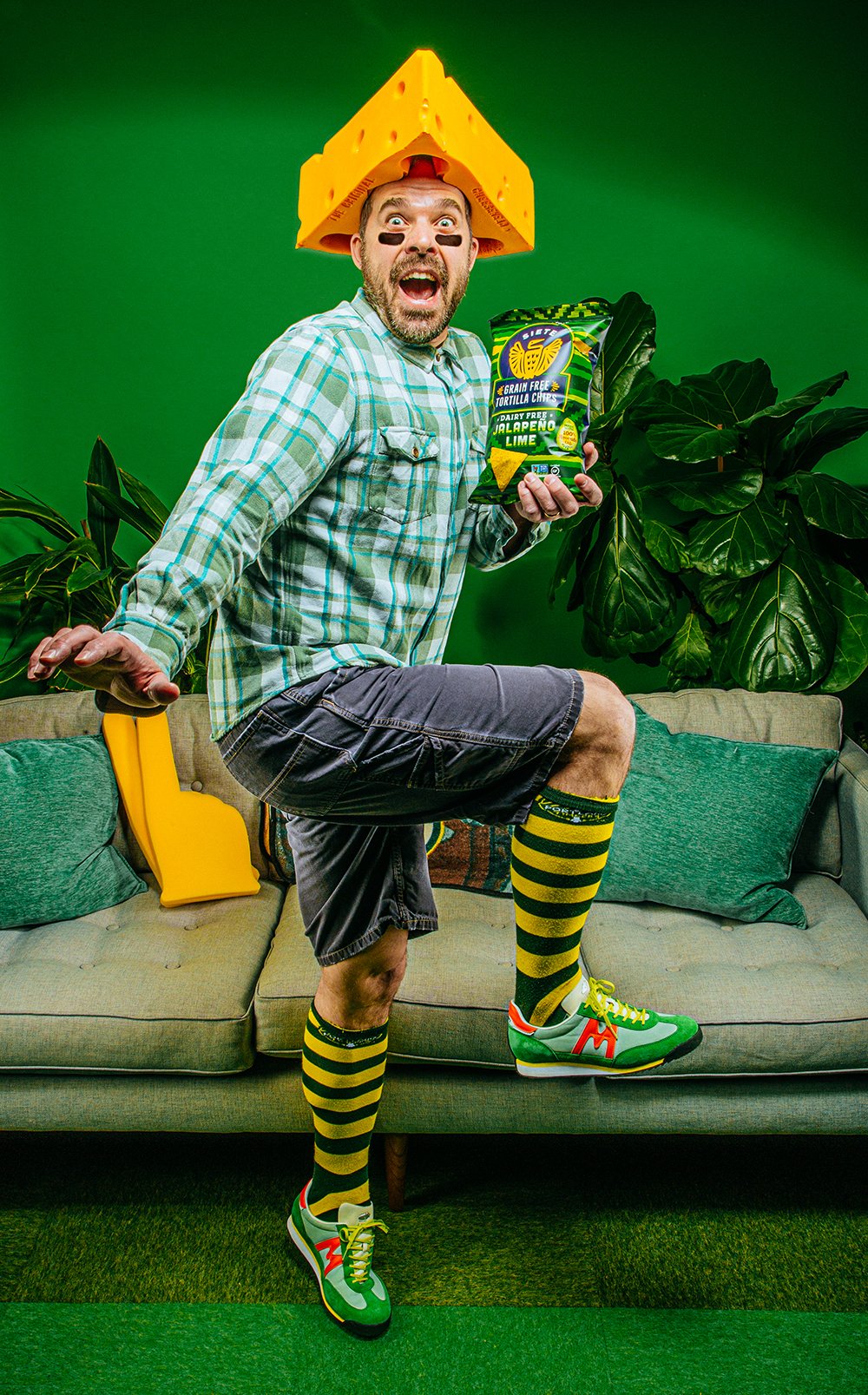 Man in cheesehead hat holding Siete jalapeno lime tortilla chips on a green couch