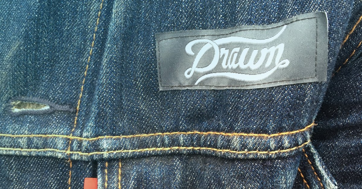 Close-up of a white "Drawn" script logo label sewn onto a dark denim jacket