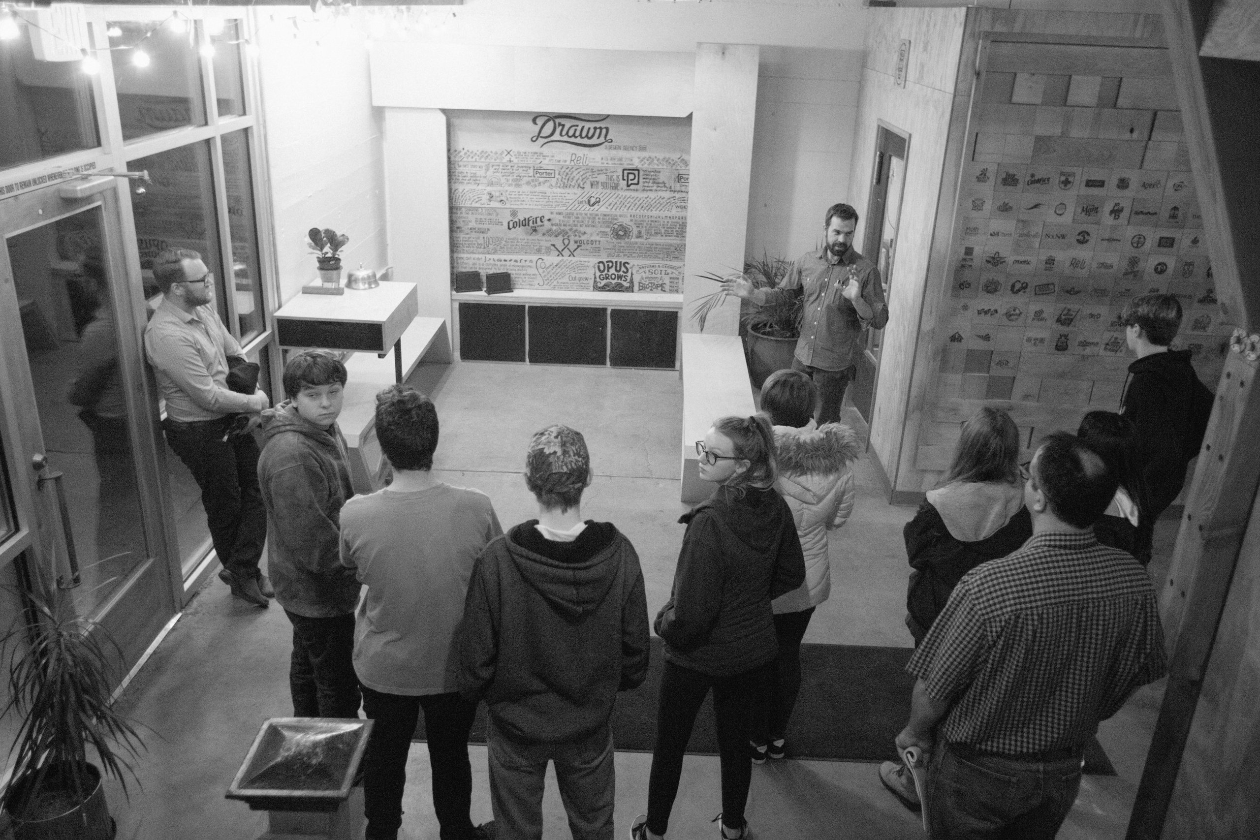 Black-and-white photo of a man addressing a group at the Drawn studio logo wall