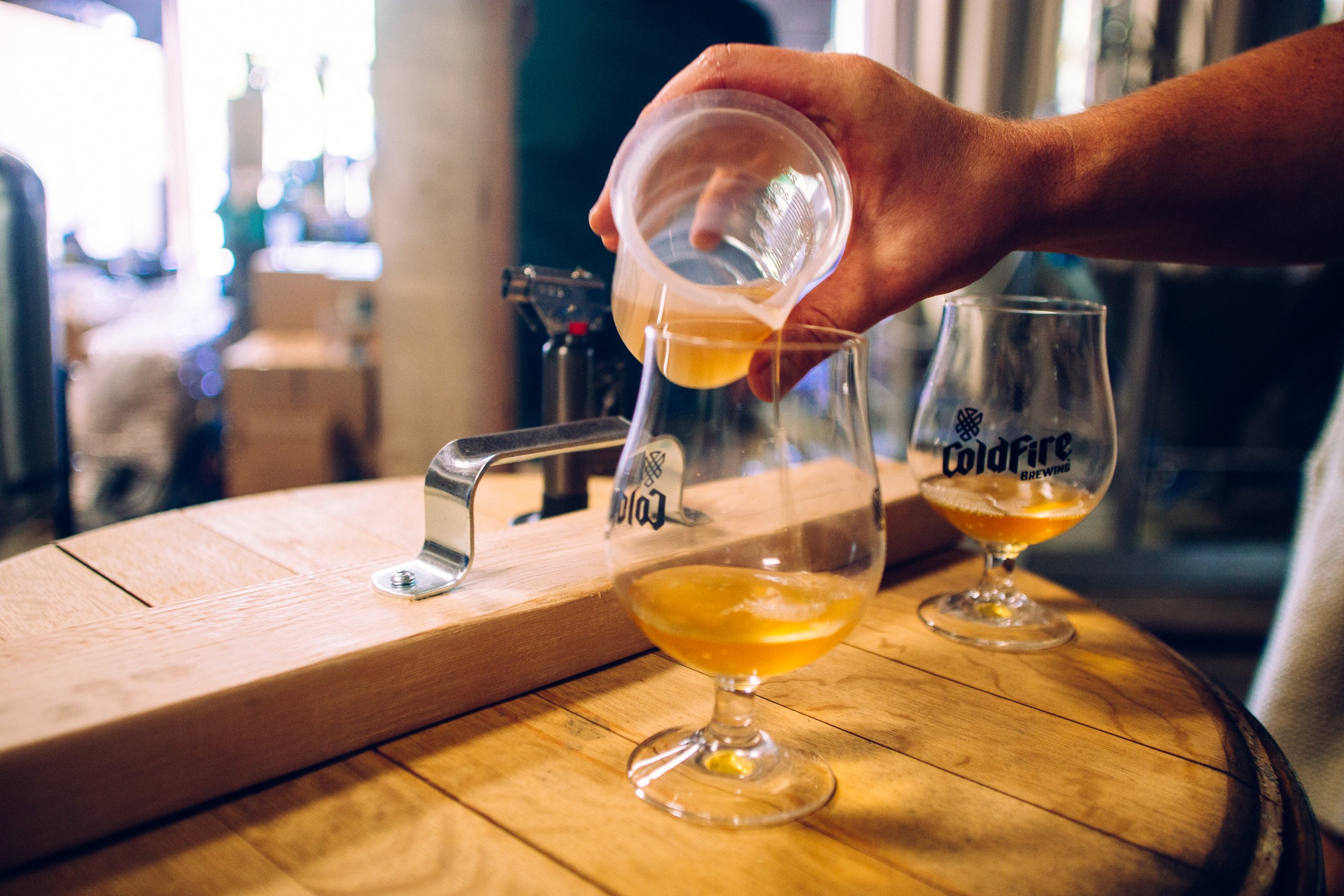 Hand pouring beer into ColdFire Brewing tasting glasses on a wooden barrel