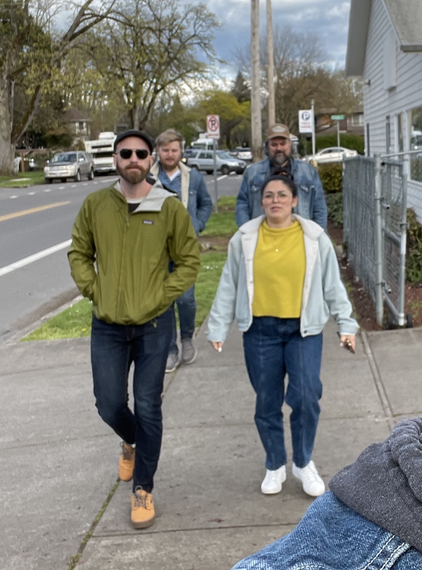 Four people walking a tree-lined sidewalk, man in olive jacket leading, woman in yellow