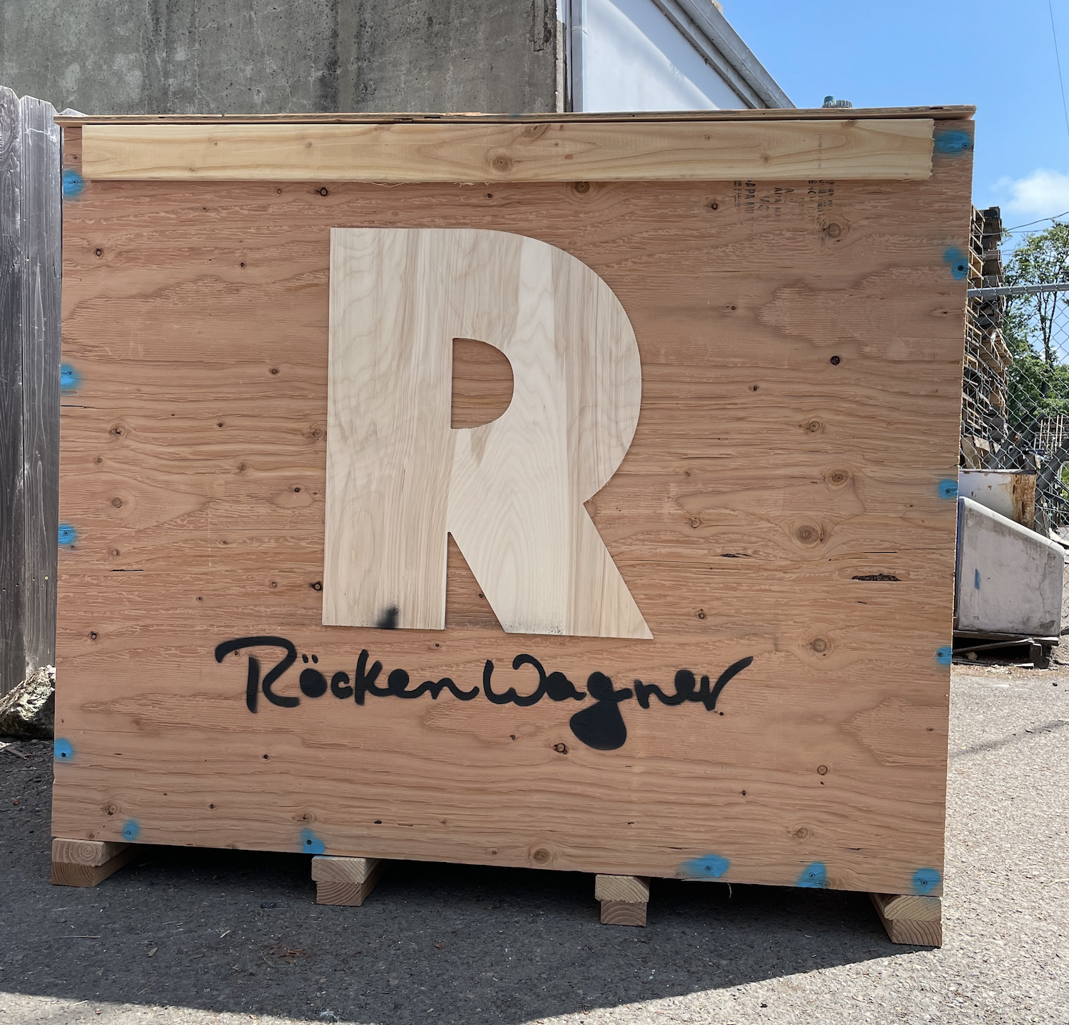 Plywood shipping crate with a cut-out R and handwritten "RockenWagner" in black paint