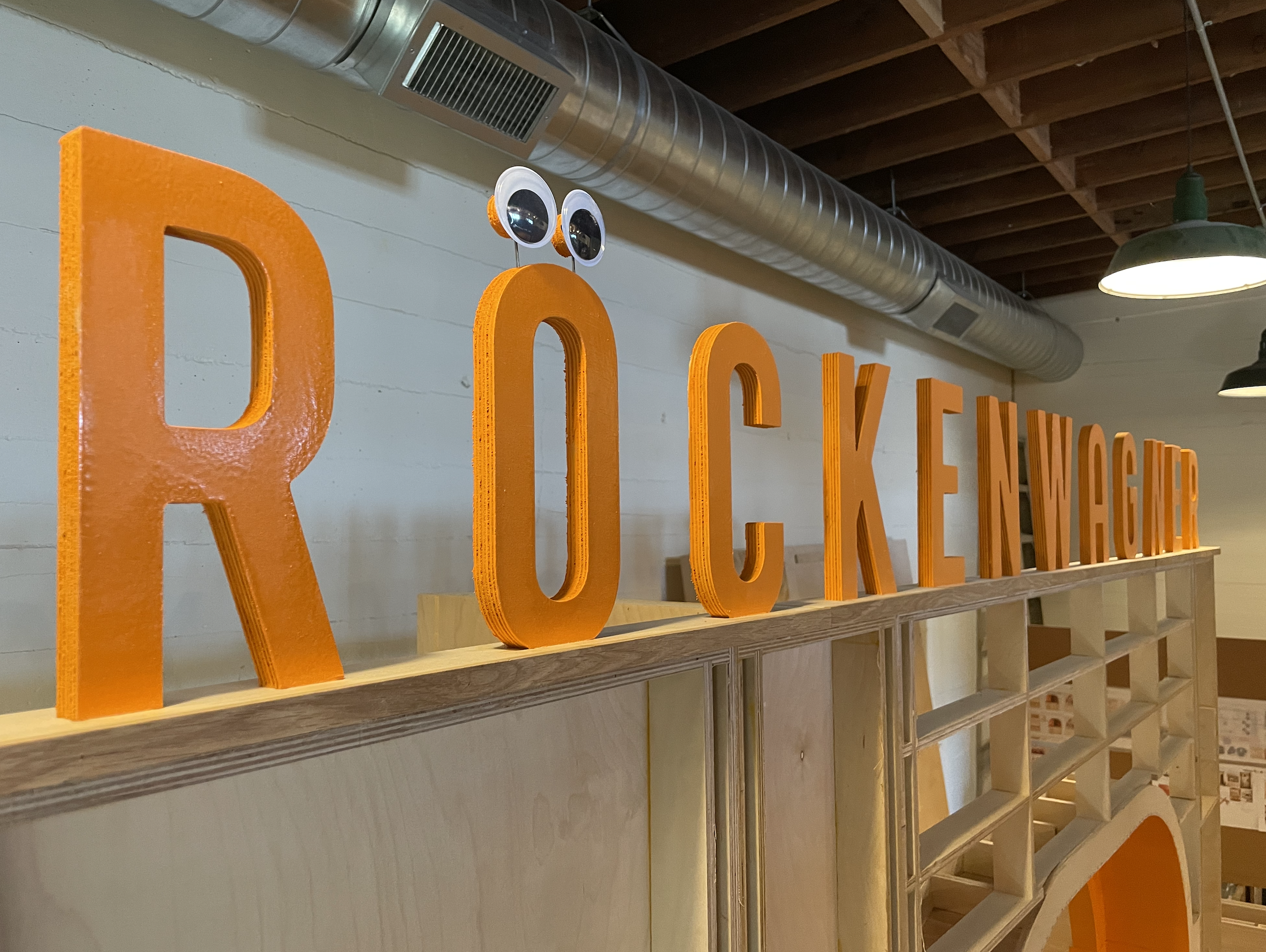 Large orange RÖCKENWAGNER cut letters on a plywood ledge, googly eyes on the Ö