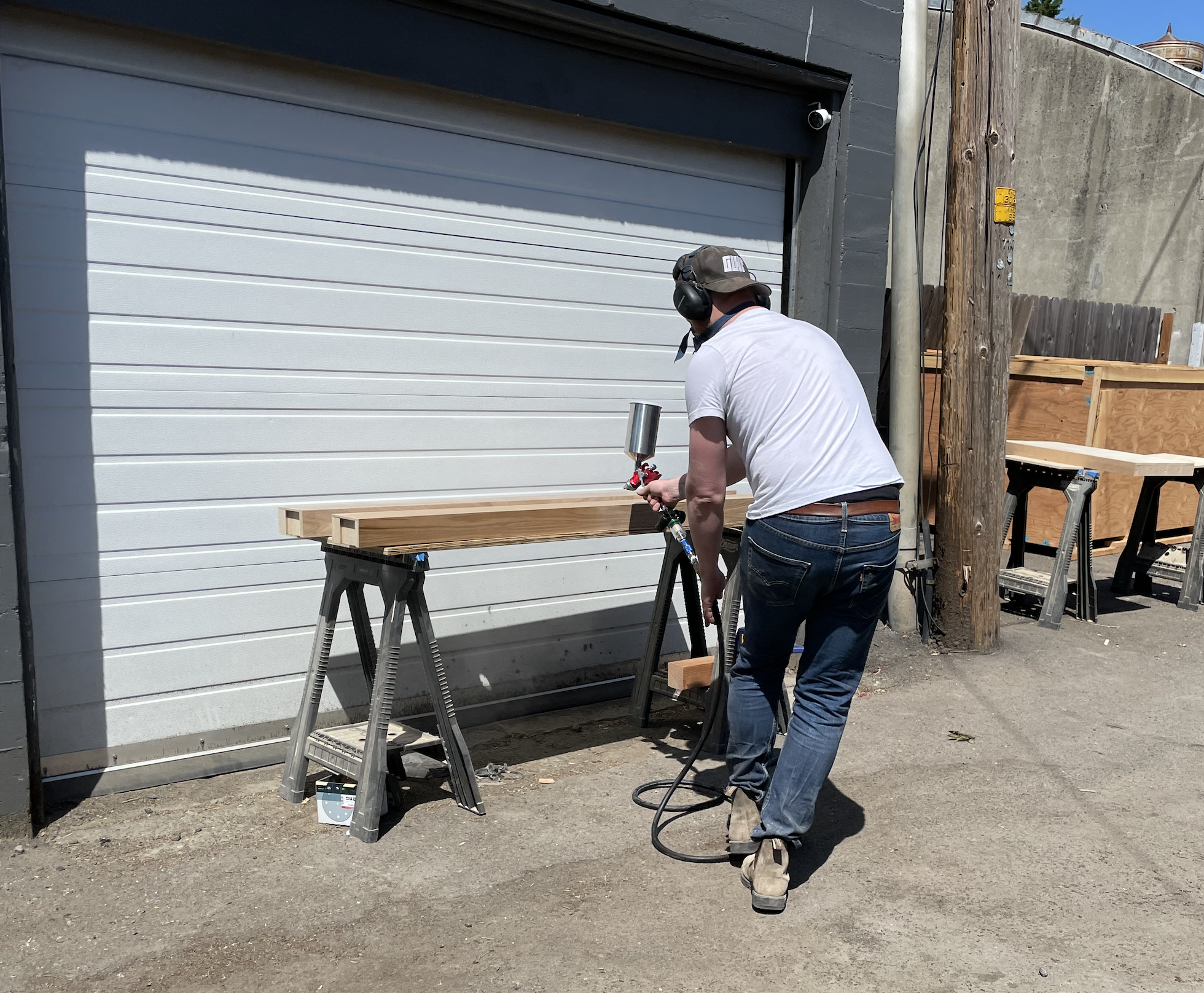 Worker in ear protection spray-finishing a wood board on sawhorses in an alley