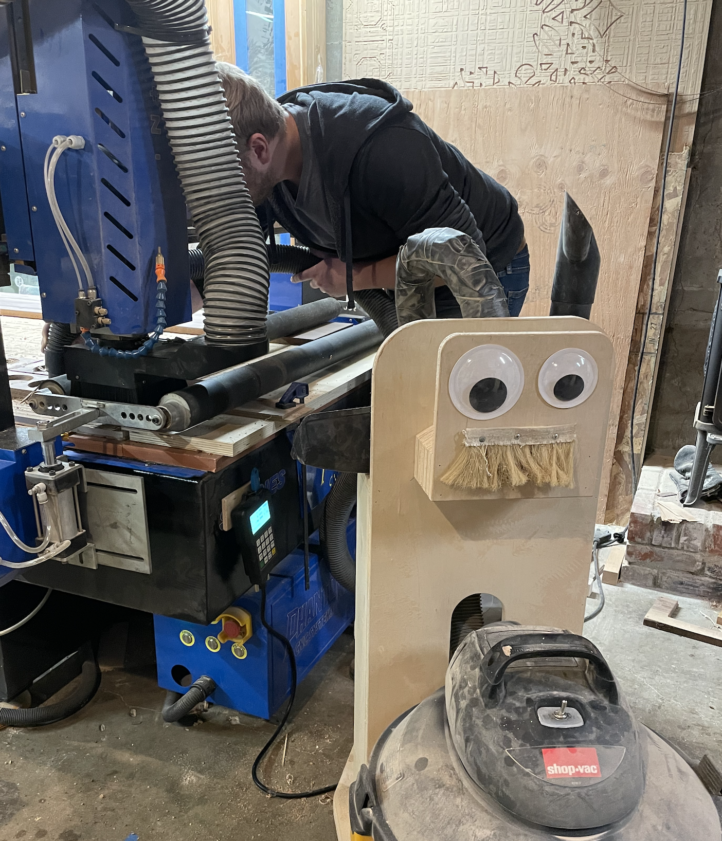 Worker at a CNC machine beside a plywood cart with googly eyes and a Shop-Vac