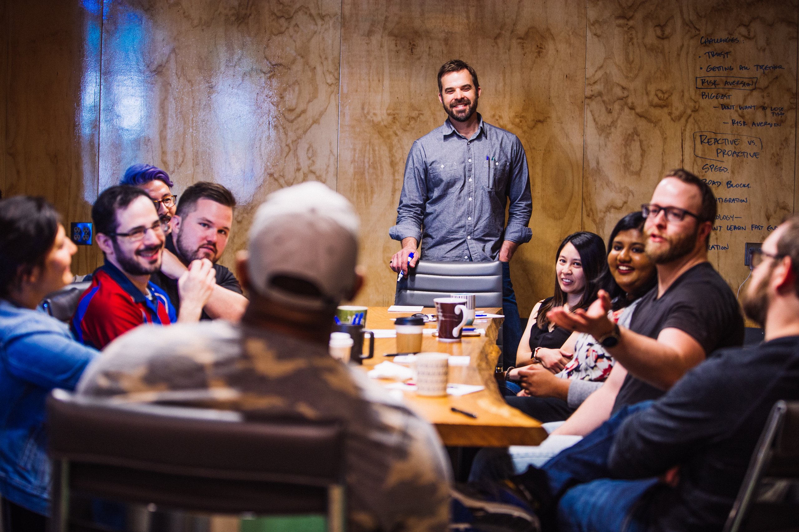 Man leading a team meeting around a table in a plywood-walled room