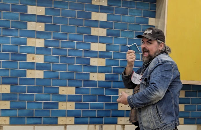 Man in denim jacket posing playfully before a blue and tan tiled wall