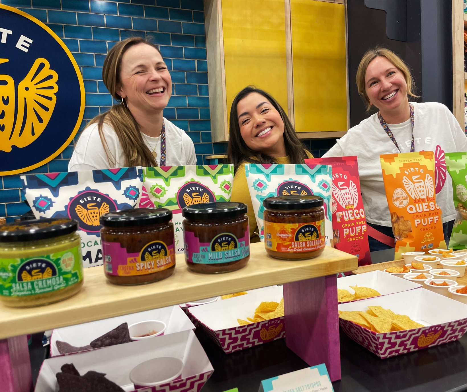 Three women smiling behind Siete salsa jars and chip bags at a booth
