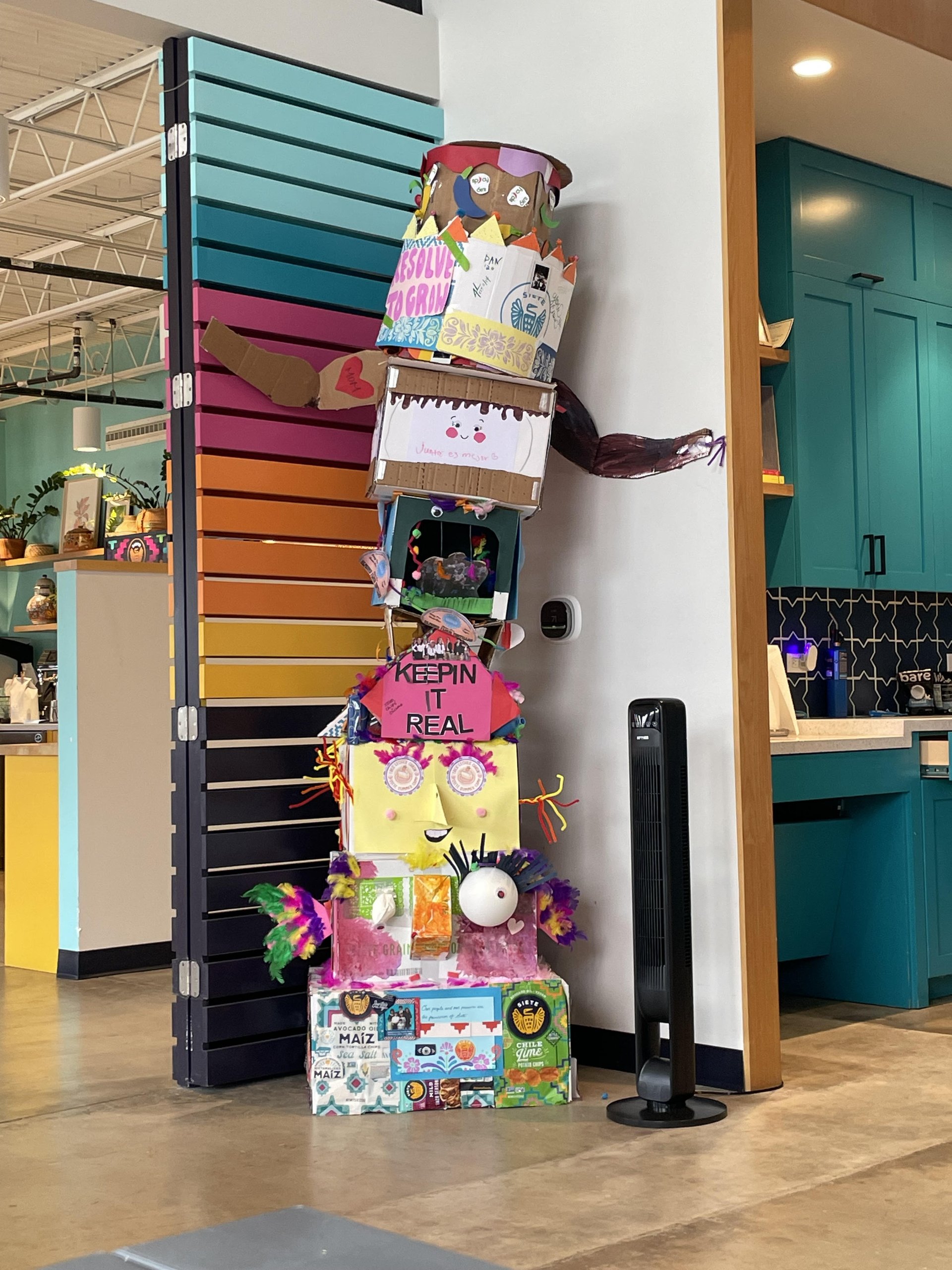 Tall cardboard totem built from Siete snack boxes reading "KEEPIN IT REAL" in a colorful office