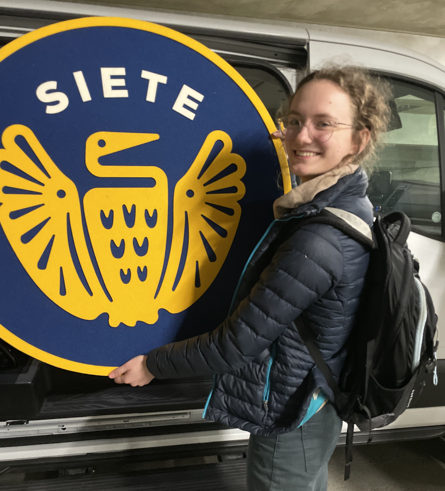 Smiling person with a backpack holding a round blue-and-yellow Siete pelican sign