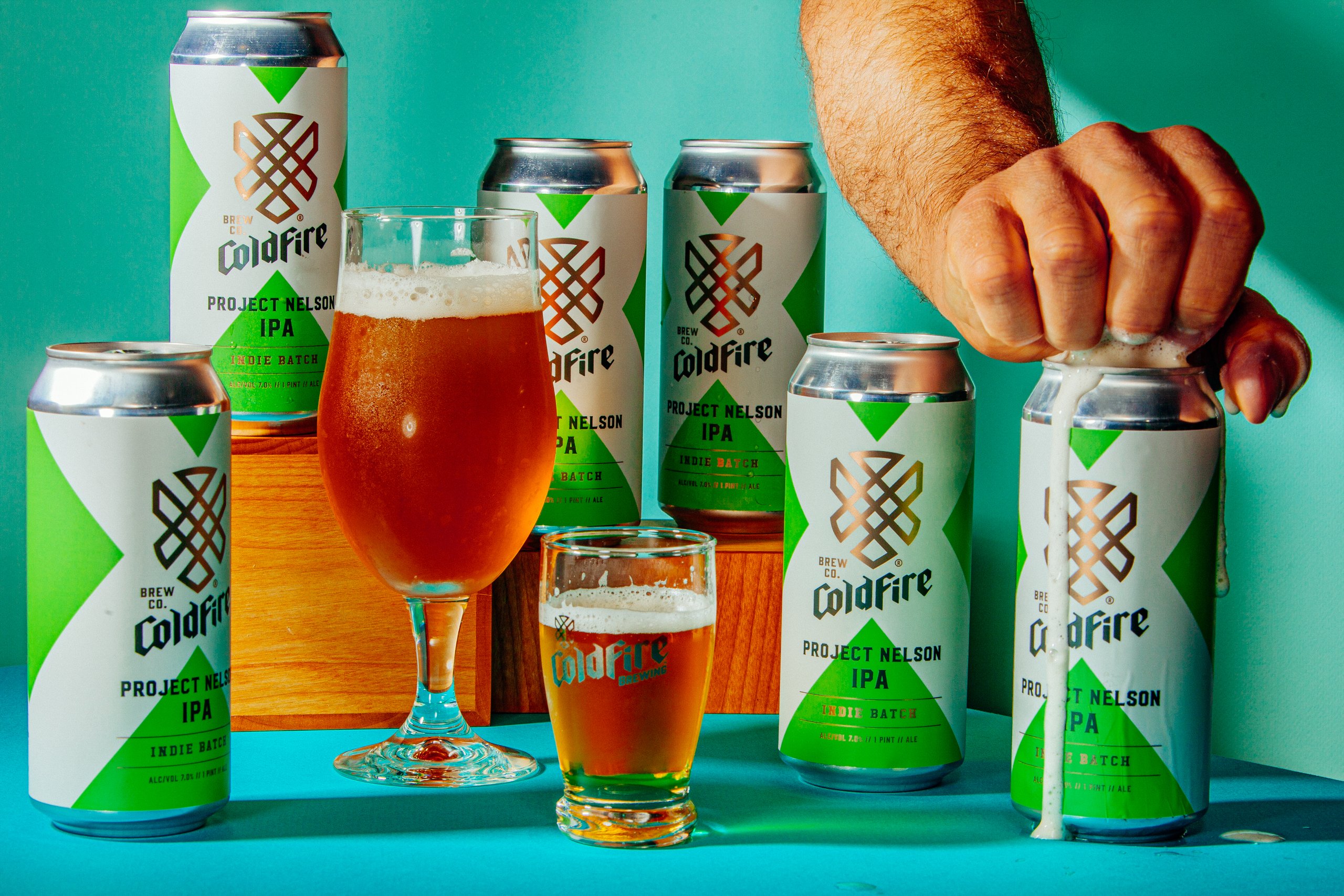 Coldfire Project Nelson IPA cans and poured glasses on teal, hand crushing one can