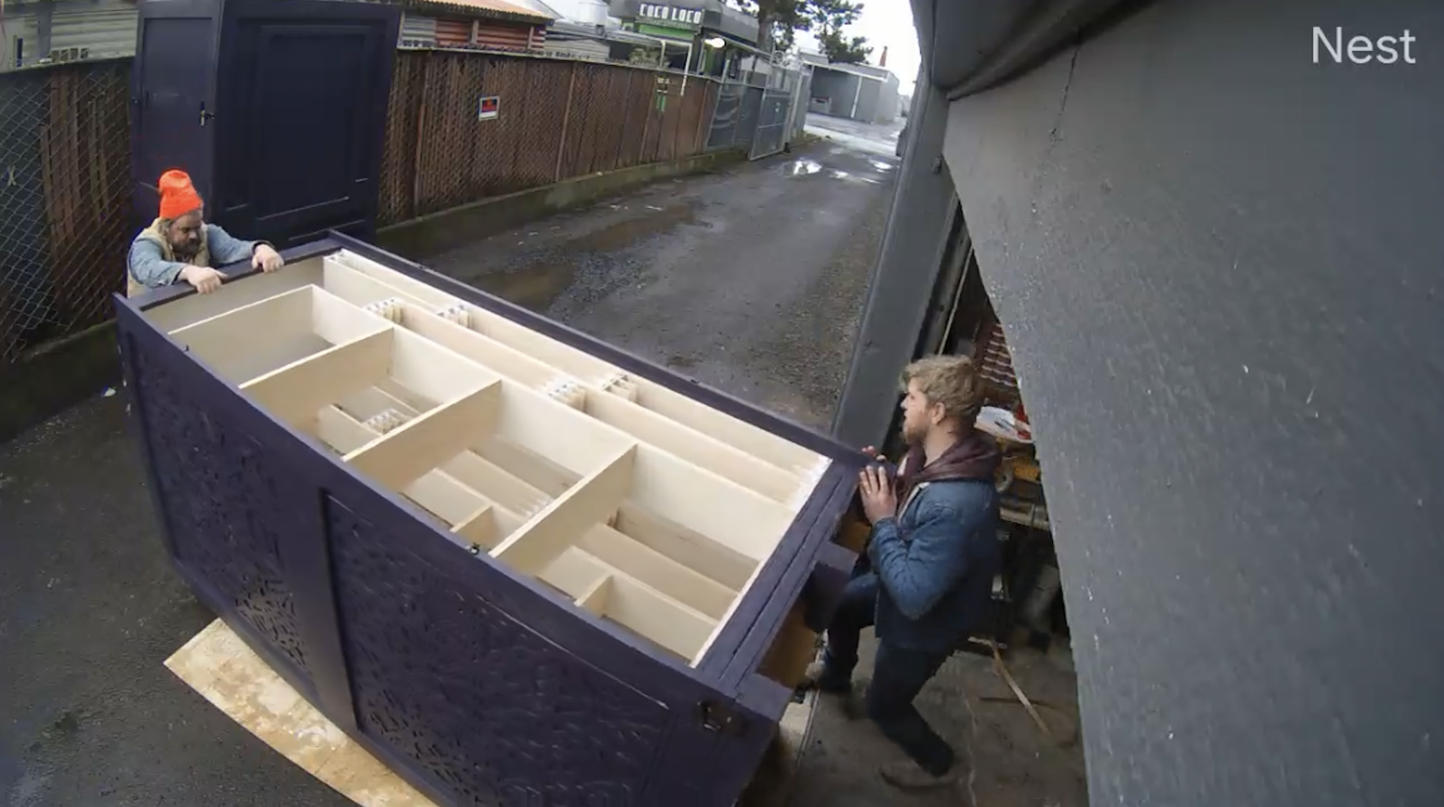 Nest camera footage of two men carrying a large purple bar cabinet down an alley