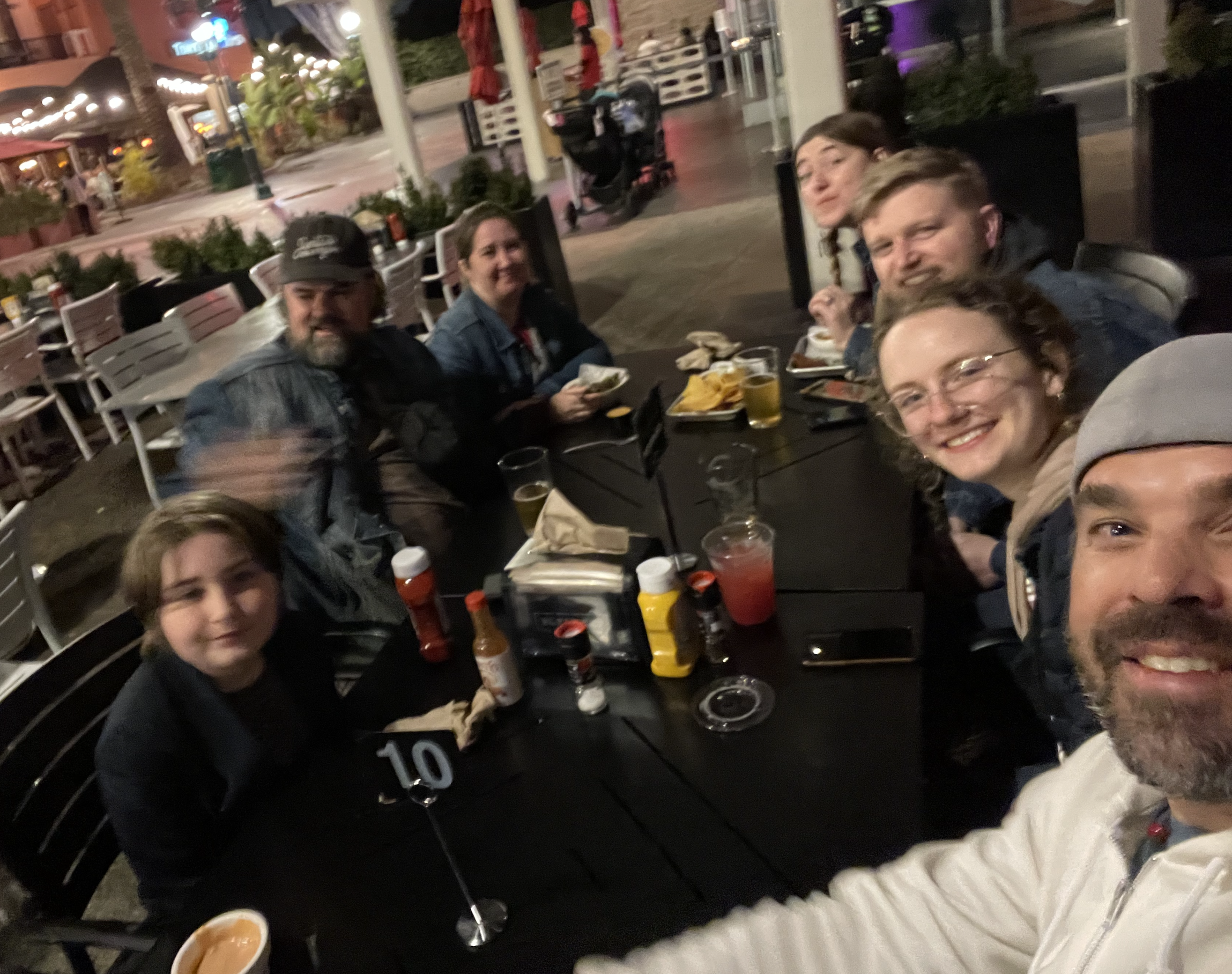 Group selfie around an outdoor restaurant table at night with food and drinks