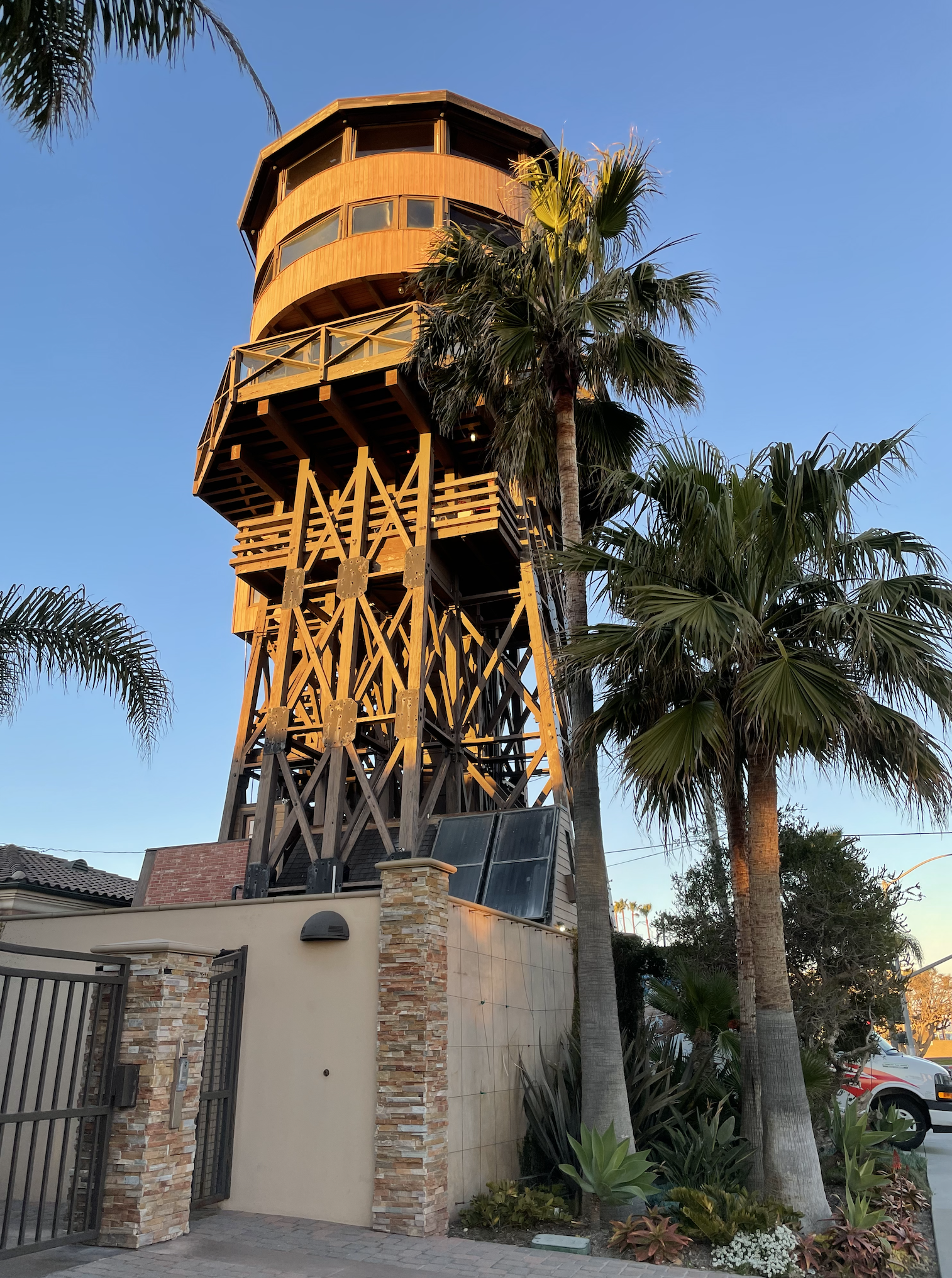 Converted wooden water-tower house glowing at dusk among tall palm trees