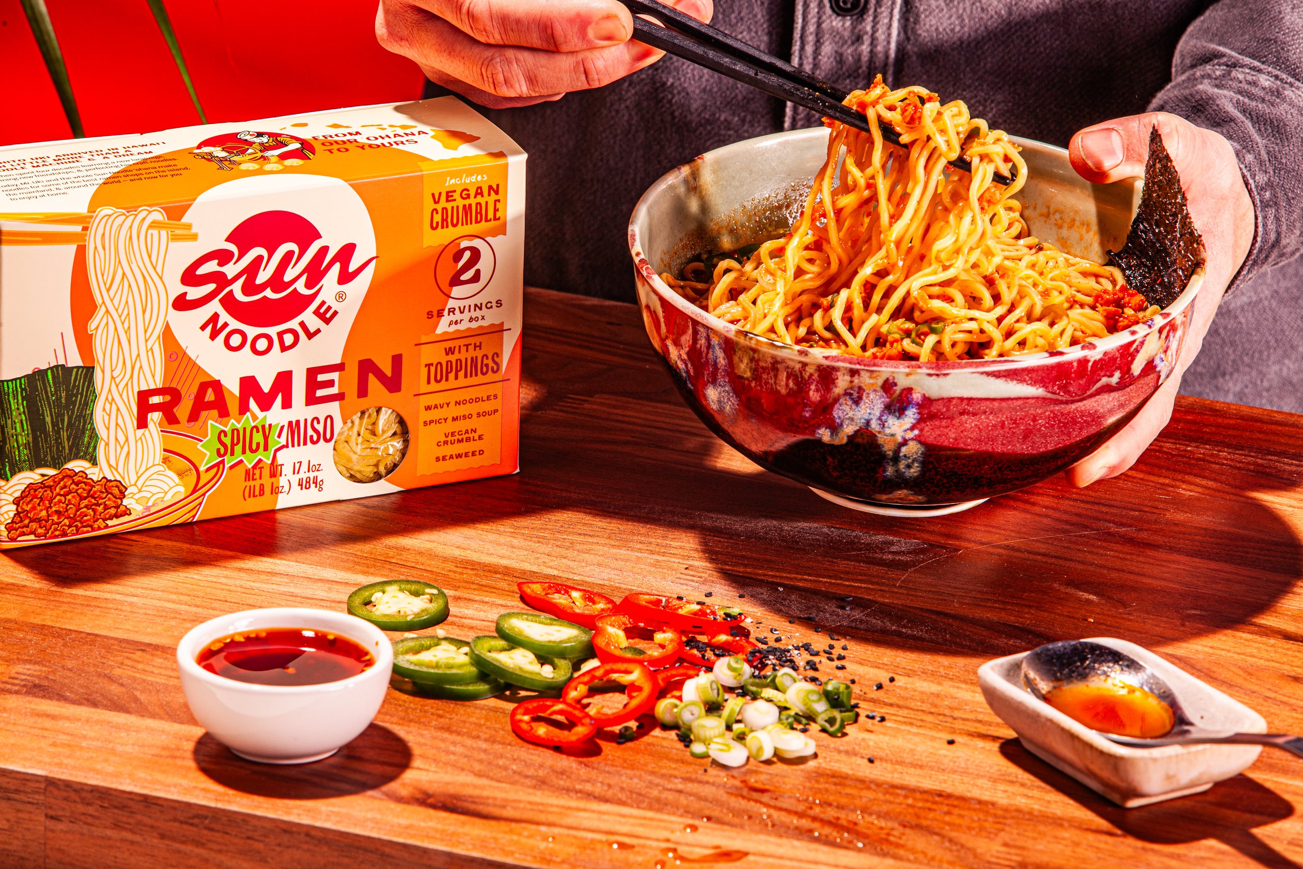 Chopsticks lifting noodles beside a Sun Noodle Spicy Miso Ramen box with sliced peppers