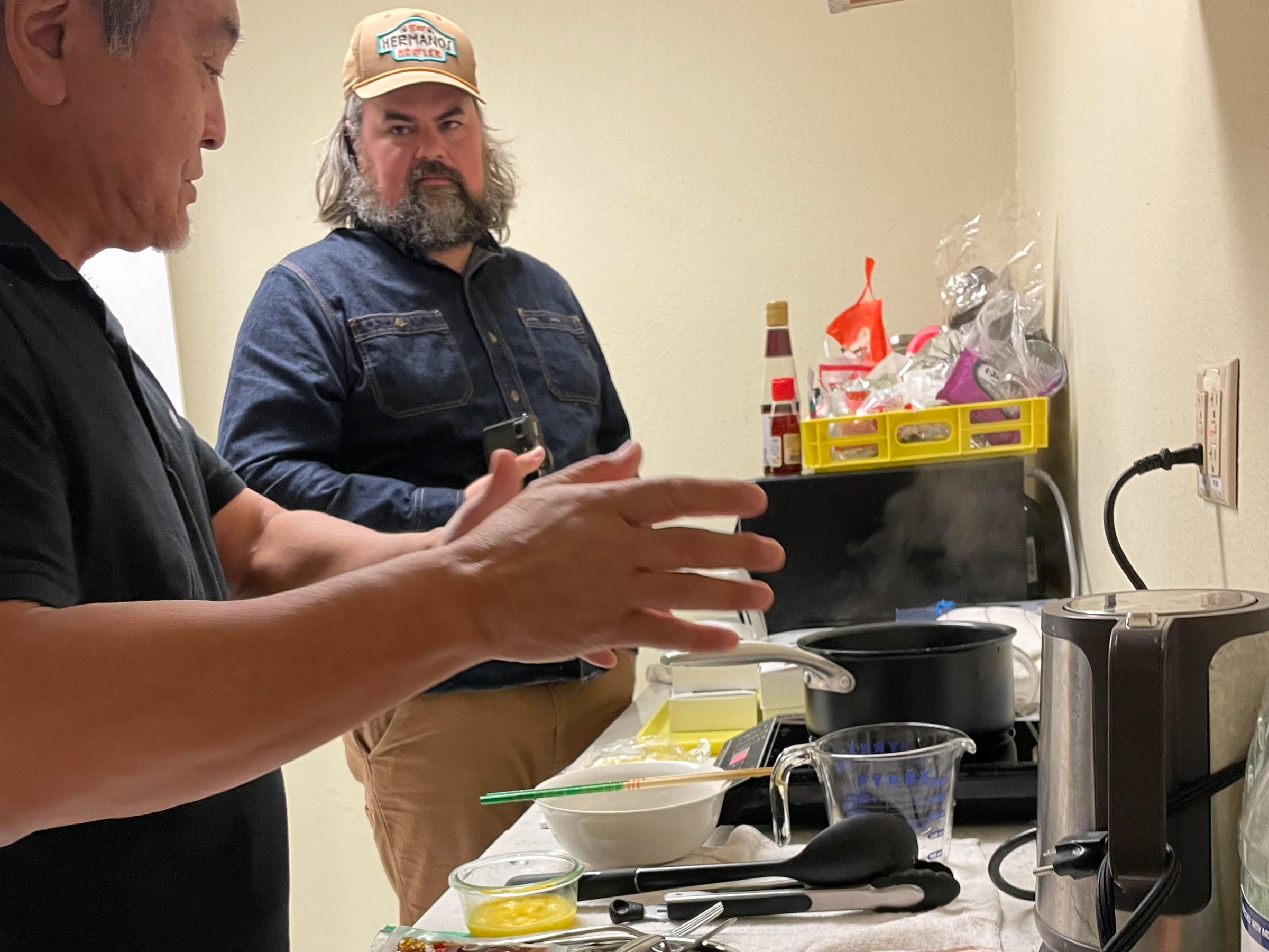 Two men in a small kitchen demonstrating ramen prep with pots and ingredients on the counter