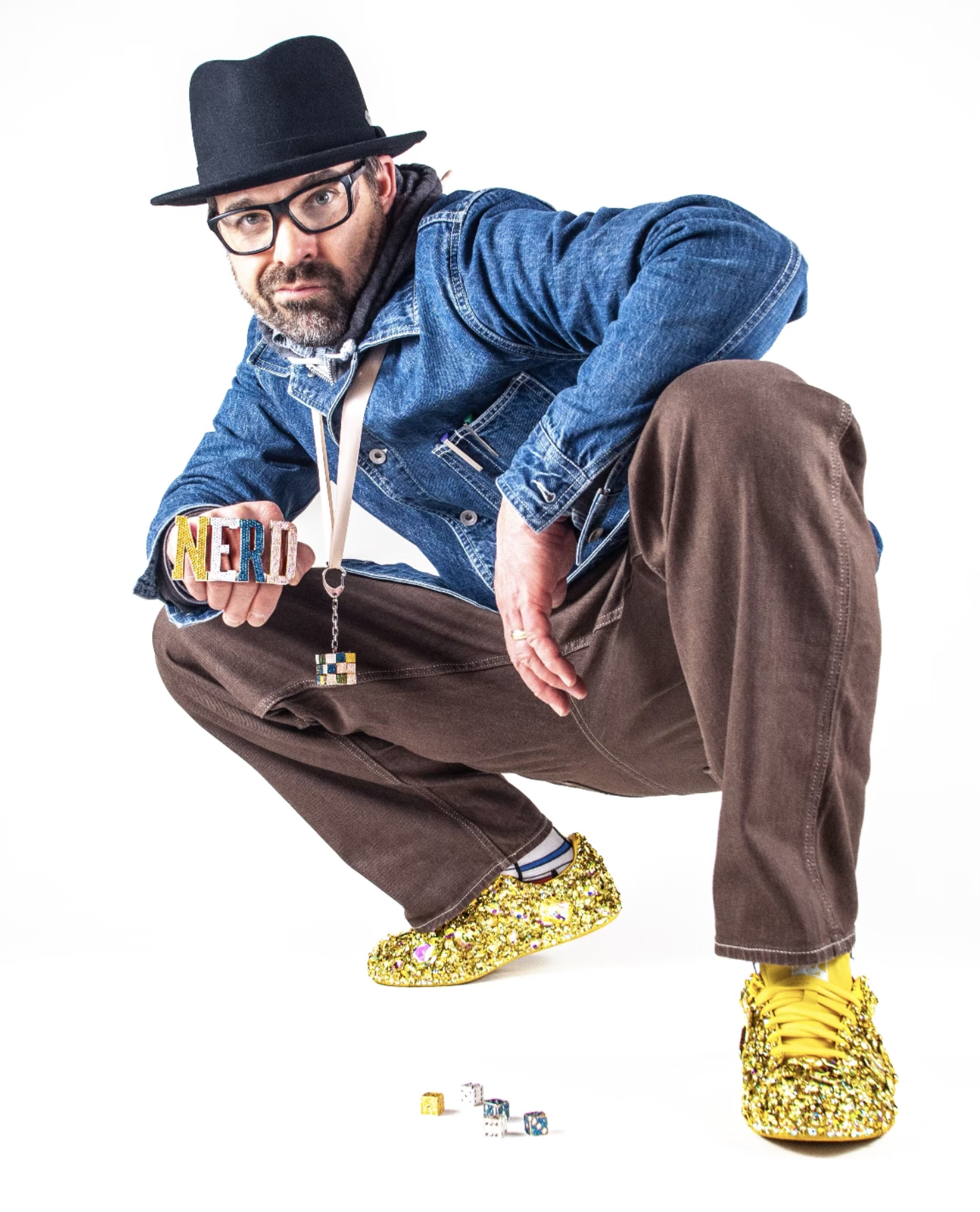 Man in fedora and glasses crouching, holding a jeweled NERD clutch in glitter sneakers