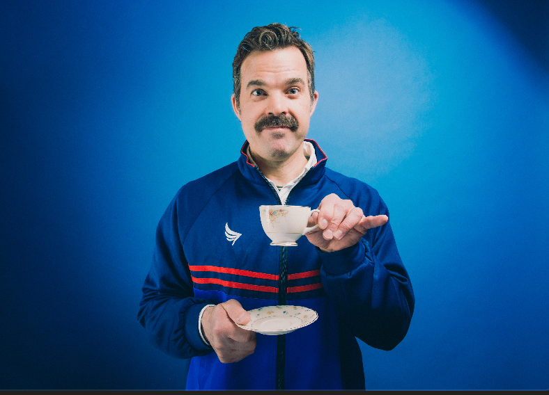 Mustachioed man in blue track jacket holding teacup and saucer, Ted Lasso costume